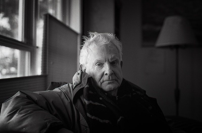 Douglas J. Penick, a black-and-white portrait by a window, in a dark jacket and scarf.